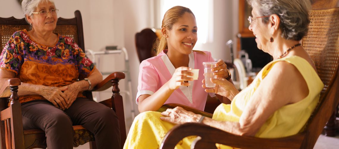 Old people in geriatric hospice: young attractive hispanic woman working as nurse helps a senior woman. She gives a water glass and prescription medicine to the aged patient