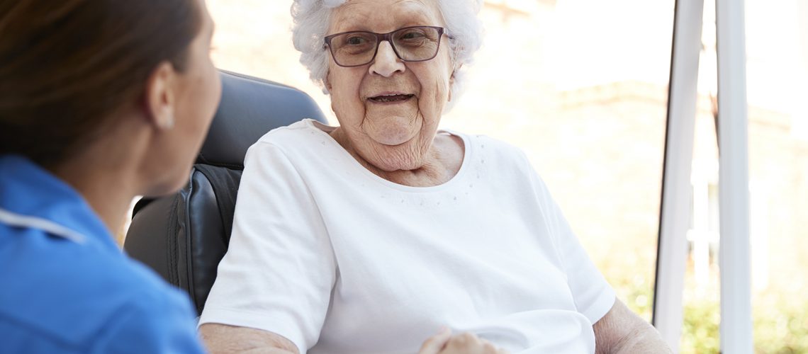 Senior Woman Sitting In Motorized Wheelchair Talking With Nurse In Retirement Home